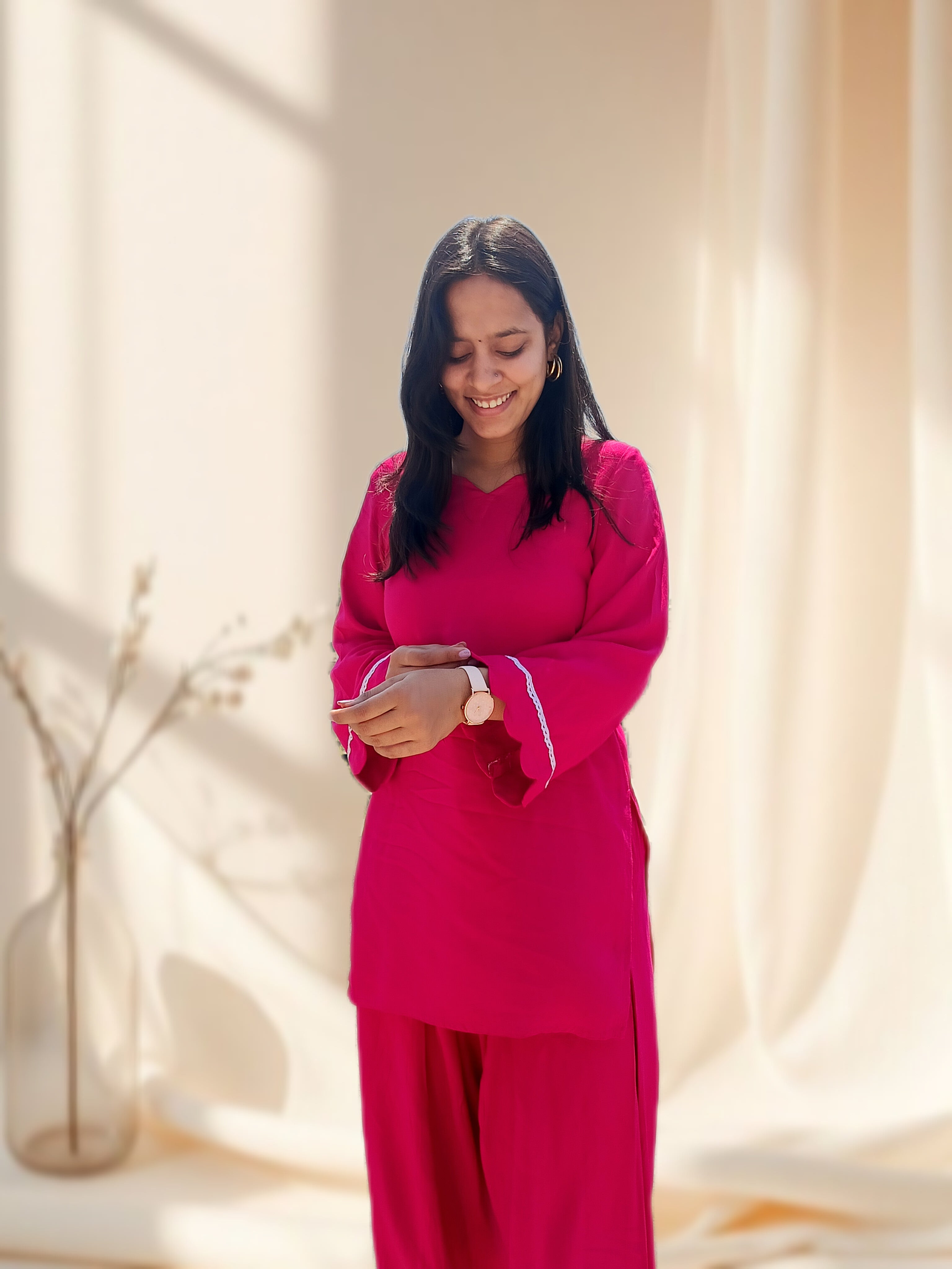 Woman in a pink outfit standing in a softly lit room with curtains.