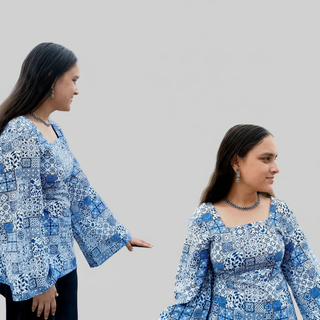 Two women in blue patterned outfits standing in front of a decorative wall with an archway.