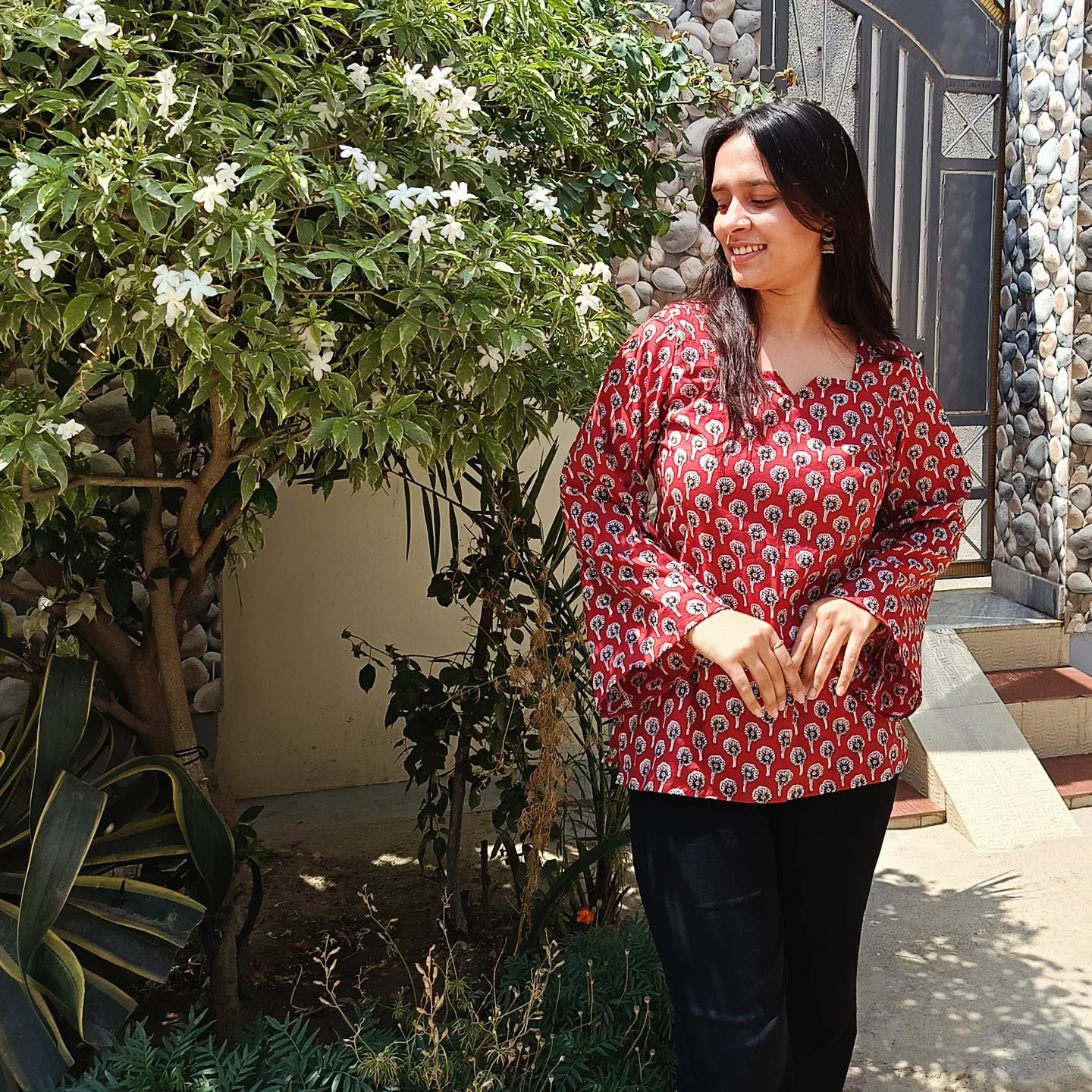 Woman in a red patterned kurti
standing outdoors near plants and a stone wall.