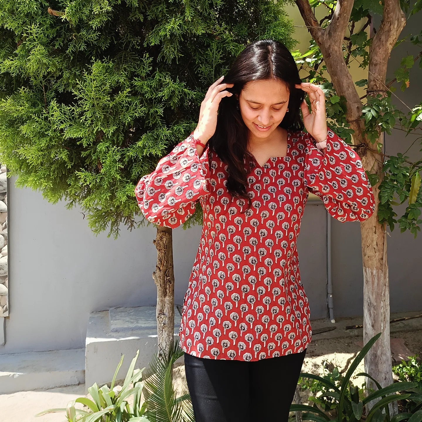 Woman wearing a red patterned kurti 
standing outdoors with greenery around.